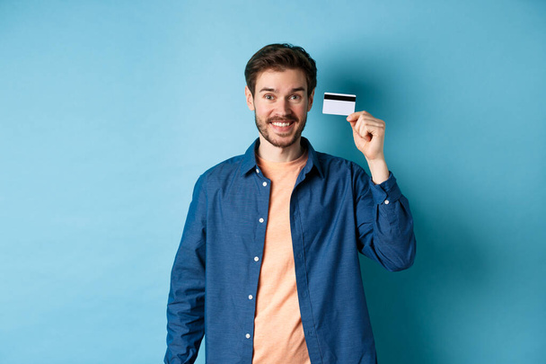 Feliz chico caucásico sonriendo, mostrando tarjeta de crédito de plástico, de pie en ropa casual sobre fondo azul - Foto, imagen