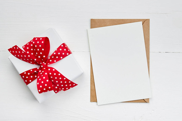 Valentines day greeting card mockup and gift box with red ribbon on white wooden background, top view, copy space - 写真・画像