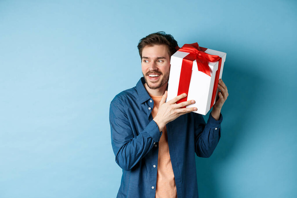 Valentines day. Smiling man guessing what inside gift box, got present from lover girlfriend, standing against blue background - Photo, Image