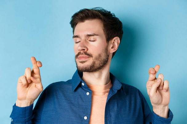 Close up of hopeful young man cross fingers for good luck and close eyes, making wish with calm expression, standing on blue background - 写真・画像