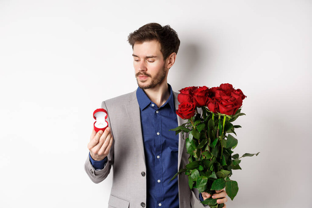 Όμορφος γενειοφόρος άντρας με κοστούμι κοιτάζει το δαχτυλίδι αρραβώνων, κάνοντας έκπληξη την ημέρα των εραστών, στέκεται με κόκκινα τριαντάφυλλα πάνω από λευκό φόντο - Φωτογραφία, εικόνα