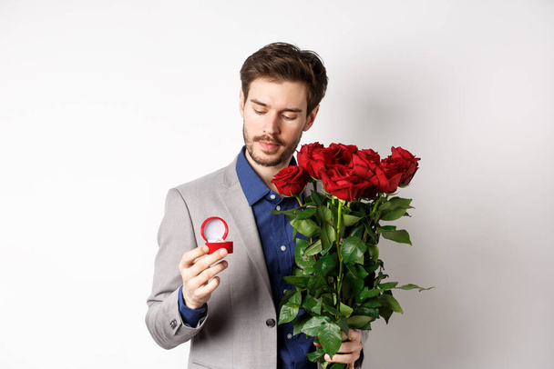 Romantikus öltönyös férfi, aki az eljegyzési gyűrűn töpreng, Valentin-napon házassági ajánlatot fog tenni, rózsacsokrot tart, fehér háttér fölött áll. - Fotó, kép