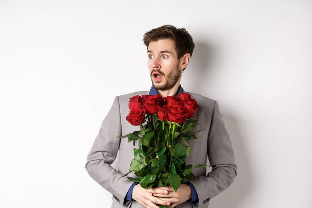 Hübscher junger Mann im Anzug, rote Rosen in der Hand, mit überraschtem und erschrockenem Gesichtsausdruck links stehend am Valentinstag vor weißem Hintergrund - Foto, Bild