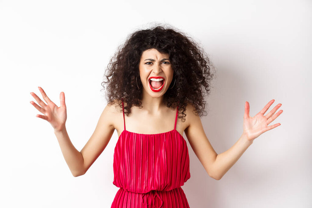 Angry young woman with curly hair, wearing red dress, screaming and having an argument, looking with hatred and anger, standing over white background - Photo, image