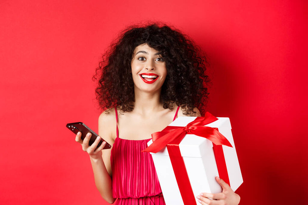 Online αγορές και ημέρα του Αγίου Βαλεντίνου. Όμορφη νεαρή γυναίκα κρατώντας smartphone και εραστές δώρο, που αναζητούν έκπληκτος και χαρούμενος στην κάμερα, κόκκινο φόντο - Φωτογραφία, εικόνα