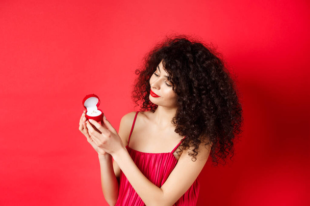 Mujer romántica de pelo rizado en vestido rojo, mirando feliz en el anillo de compromiso, convertirse en una novia, de pie en el fondo del estudio - Foto, imagen