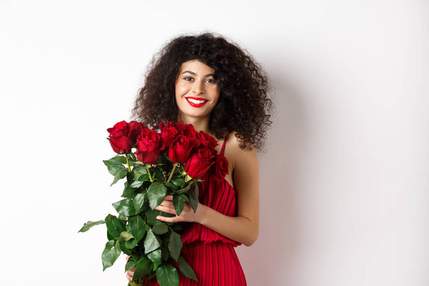 Прекрасна каукасійка в червоному вечірньому вбранні отримує букет квітів. Дівчинка з червоними трояндами посміхається перед камерою, має романтичне побачення в день кохання Валентина, біле тло - Фото, зображення