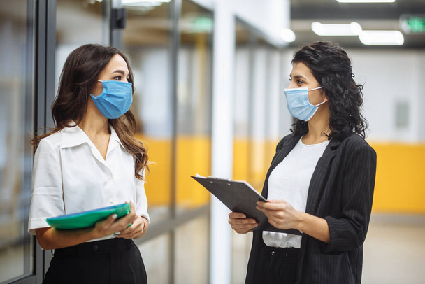 Two businesswomen discuss working matters at the office wearing medical sterile masks. Working during coronavirus pandemic quarantine. New normal, health care and safety measures concept - Photo, Image