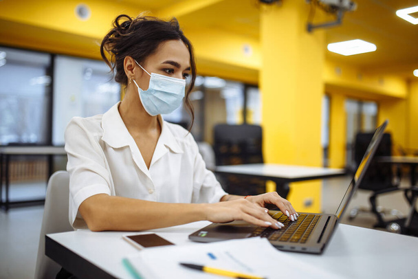 Ritratto di una donna che lavora su un computer portatile alla scrivania di un ufficio, indossa una maschera medica e rimane al sicuro durante l'epidemia di pandemia di Covid-19. Concetto di assistenza sanitaria e autoigiene - Foto, immagini