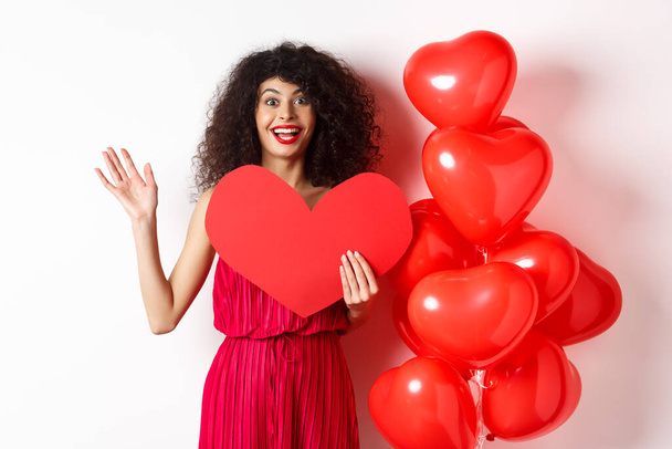 Valentines day and love concept. Cheerful young woman in elegant red dress, standing near romantic balloons and holding big red heart cutout, waving hand to say hi, waiting for date - Foto, Imagen