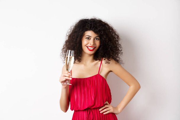 brutale jonge vrouw in elegante rode jurk, poseren met glas champagne op witte achtergrond, met romantische date op Valentijnsdag - Foto, afbeelding