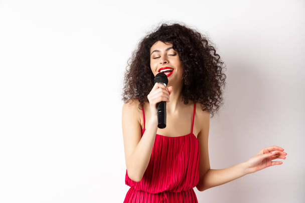 Szczęśliwa elegancka kobieta w czerwonej sukience wykonująca z mikrofonem, śpiewająca karaoke i uśmiechnięta, stojąca nad białym tłem - Zdjęcie, obraz