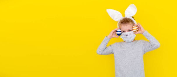 Banner copyspace Boy in rabbit bunny ears on head and protective mask with colored eggs on yellow studio background. Cheerful crazy smiling happy child. Covid easter kid - Фото, изображение