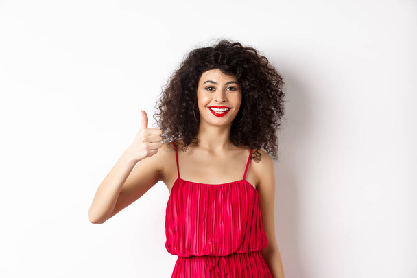 Cheerful woman with red lips and elegant dress, showing thumb up and smiling, recommending product, standing over white background - Photo, Image