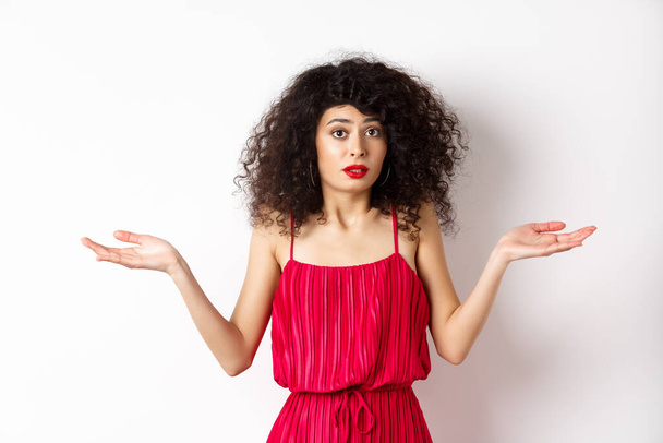 Confused lady in red dress shrugging shoulders, spread hands sideways and looking puzzled, know nothing, cant understand, standing over white background - Foto, Imagen