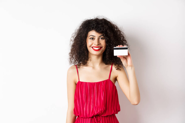 Kırmızı elbiseli ve makyajlı güzel bir kadın, plastik kredi kartı gösteriyor ve gülümsüyor, öneri sunuyor, beyaz arka planda duruyor. - Fotoğraf, Görsel