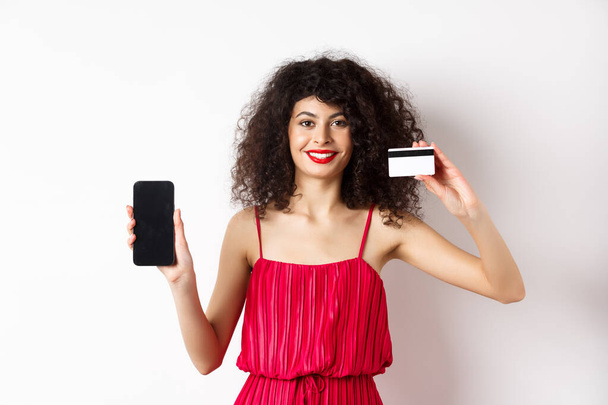 Online-Einkaufskonzept. Elegantes weibliches Model mit lockigem Haar, trägt rotes Kleid, zeigt Plastikkreditkarte mit leerem Handy-Bildschirm, steht auf weißem Hintergrund - Foto, Bild