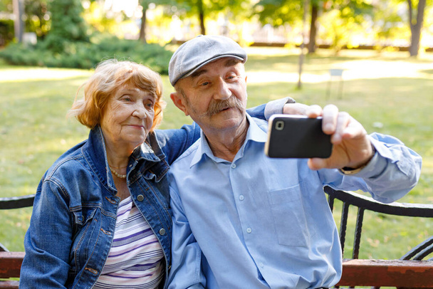 Ein älteres Ehepaar posiert in einem Park für ein Selfie. Technik, Kommunikation und Menschen - ältere Ehepartner mit Smartphone. Der Alltag eines älteren Menschen. - Foto, Bild