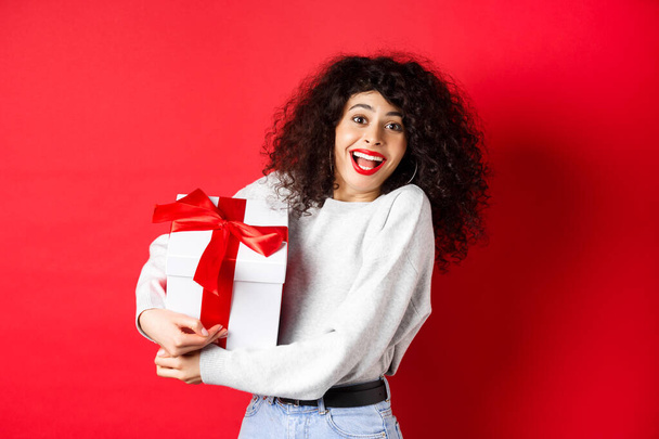 Valentines day. Happy girlfriend holding gift box from secret admirer, looking romantic at lover, receive present, standing on red background - Fotó, kép