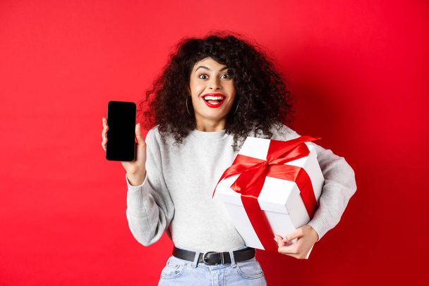 Ημέρα του Αγίου Βαλεντίνου και εραστών. Ενθουσιασμένη χαμογελαστή γυναίκα με σγουρά σκούρα μαλλιά, που δείχνει το smartphone άδειο οθόνη και κρατώντας δώρο έκπληξη στις διακοπές, που δείχνει online promo, κόκκινο φόντο - Φωτογραφία, εικόνα