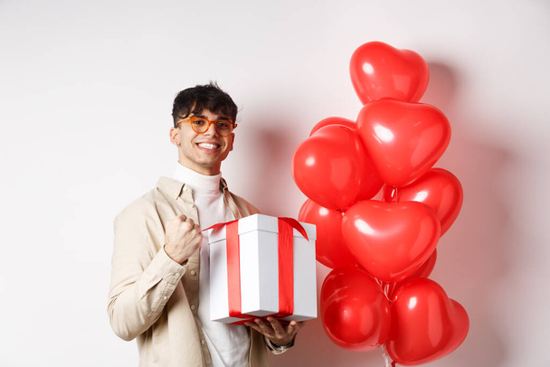 Valentines day and romance concept. Happy and confident boyfriend prepare gift for lover, saying yes and smiling, holding romantic gift, standing near red hearts balloons - Valokuva, kuva
