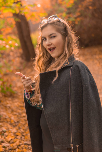 A cheerful young girl in a black cloak stands in chameleon glasses in autumn. Yellow forest, green grass, blonde. well-being concept with charismatic appearance - Foto, Imagen