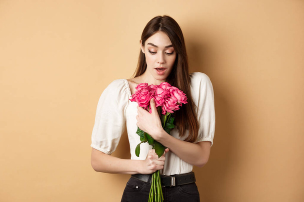 Walentynki. Zaskoczona dziewczyna otrzymać prezent od kochanka na dzień, patrząc zdumiony pięknym bukietem kwiatów, trzymając róże na beżowym tle - Zdjęcie, obraz