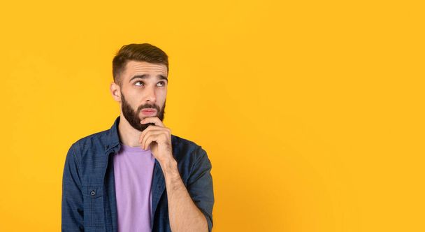 Dreamy Caucasian guy thinking over something, touching his chin on orange background, banner design with copy space - Фото, изображение