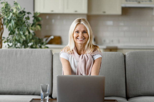 Attractive middle aged caucasian blonde sits at home in living room, using laptop, looks at camera and smiling happily while shopping online, working or studying remotely - Foto, imagen