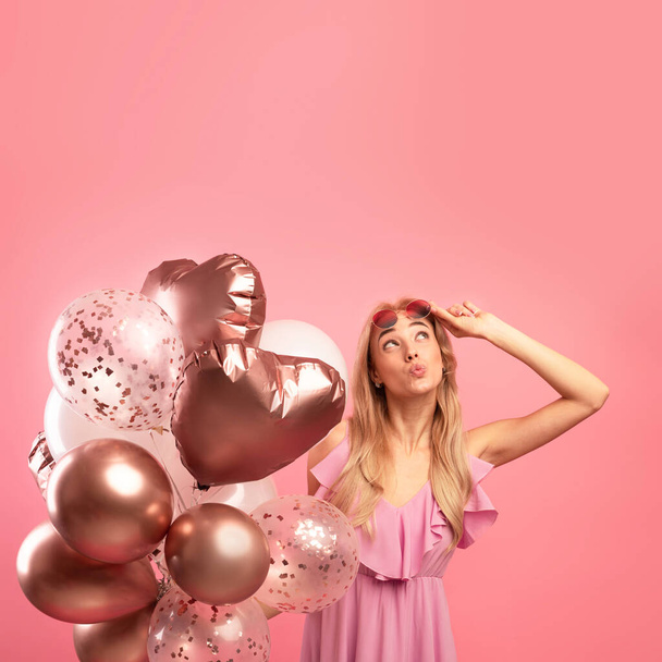 Holiday sale concept. Cute blonde woman holding bunch of balloons, pursing her lips and looking upwards at copy space - Fotó, kép