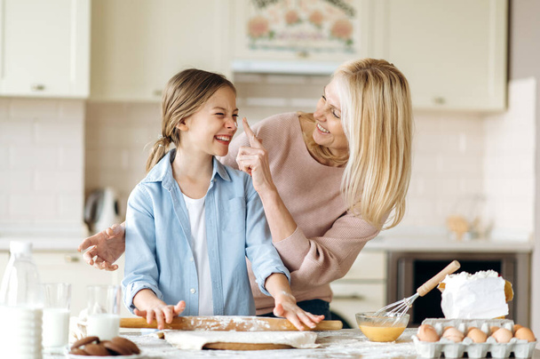 Cheerful mom or grandma and her pretty little daughter or granddaughter spend time together at the kitchen. They cook together a pie, indulge in flour, laugh and have fun - Photo, Image