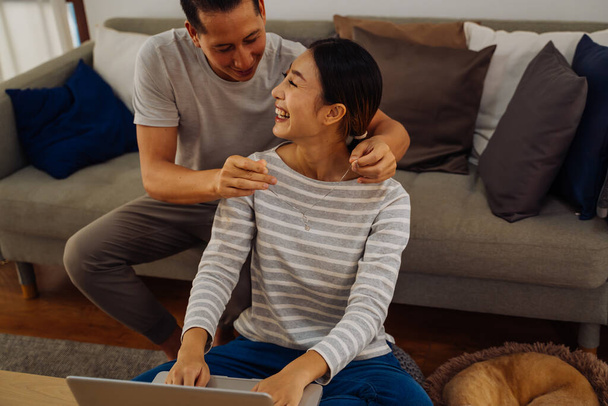 Young man giving his girlfriend a necklace as present. Asian woman getting surprised by her boyfriend with marriage proposal while working on laptop at home - Fotó, kép