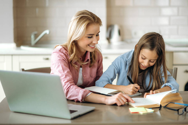 Learning at home. Caring mom helps her beloved daughter do her homework, they sit at the table at home, using a laptop, books and a notebook. Homeschooling - Zdjęcie, obraz