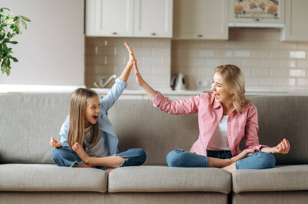 Joyful caucasian mom and daughter are sitting on the sofa at home in a lotus position, dressed in casual stylish clothes, giving high five to each other, spending time together and enjoying meditating - Fotó, kép
