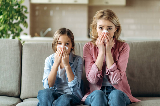 風邪をひいた悲しい白人の母親と娘は、ナプキン、くしゃみ、咳、鼻水でソファの上に家に座っています。風邪、インフルエンザ、病気 - 写真・画像
