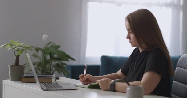 estudiante mujer está viendo conferencia por Internet y escribir notas en el cuaderno, sentado en la mesa con el ordenador portátil y mirando en la pantalla - Metraje, vídeo