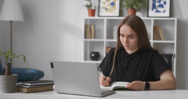 young woman is reading information on screen of laptop and writing in notebook, learning in home, distant education, online course - Footage, Video