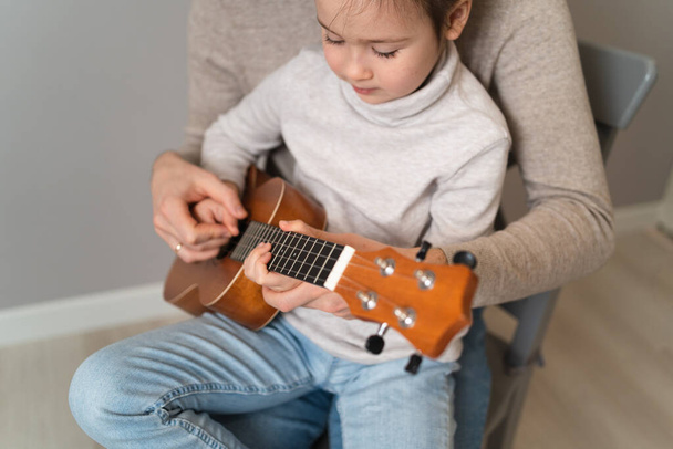 Ο μπαμπάς παίζει κιθάρα με την κόρη του. Το παιδί μαθαίνει να παίζει ένα μουσικό όργανο με έναν δάσκαλο. Μουσικό ντουέτο ενός πατέρα με ένα παιδί. - Φωτογραφία, εικόνα