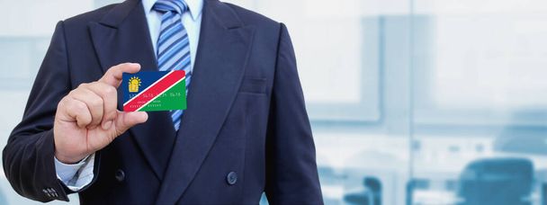 Imagen recortada del hombre de negocios sosteniendo una tarjeta de crédito de plástico con la bandera impresa de Namibia. Fondo borroso. - Foto, imagen