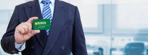 Cropped image of businessman holding plastic credit card with printed flag of Saudi Arabia. Background blurred. - Photo, Image