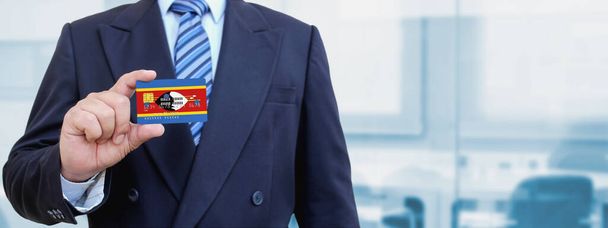 Обрезанное изображение бизнесмена, держащего пластиковую кредитную карту с напечатанным флагом Свазиленда. Фон размыт. - Фото, изображение