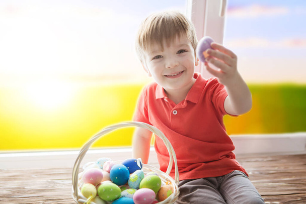 Cheerful smiling boy holding basket full of colorful easter eggs and sitting on the windowsill against the background of a spring green field at sunset or dawn - Foto, afbeelding