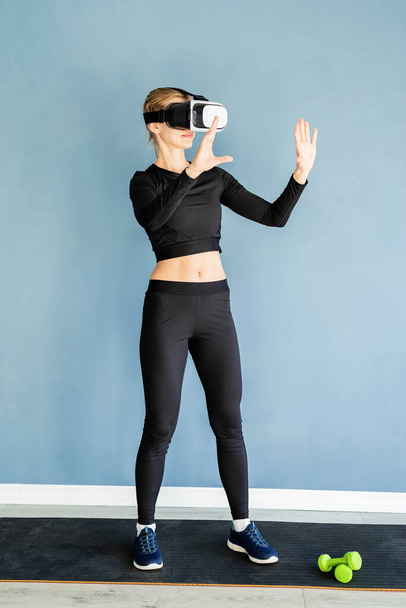 Fitness, sport and technology. Young athletic woman wearing virtual reality glasses standing at fitness mat using VR interactive menu - Fotografie, Obrázek