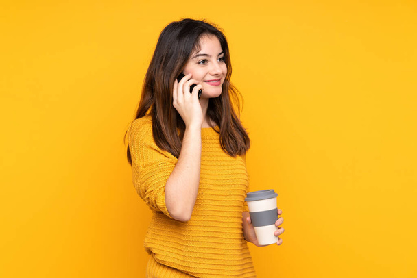 Young caucasian woman isolated on yellow background holding coffee to take away and a mobile - Foto, Imagem