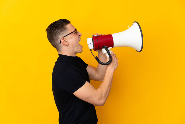 Young caucasian man isolated on yellow background shouting through a megaphone to announce something in lateral position - Fotó, kép