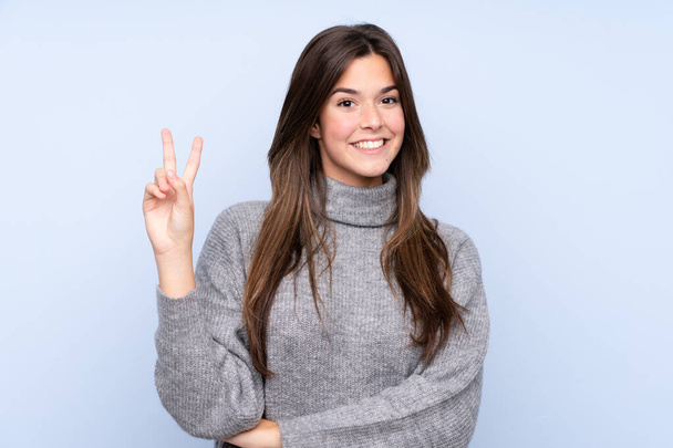 Teenager Brazilian girl over isolated blue background smiling and showing victory sign - Photo, image