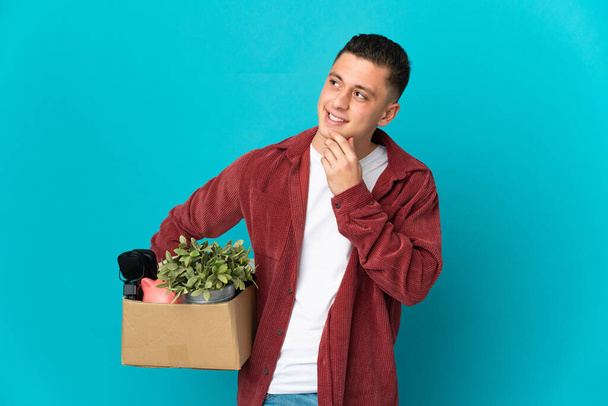 Νεαρός καυκάσιος άντρας κάνει μια κίνηση ενώ μαζεύει ένα κουτί γεμάτο πράγματα απομονωμένα σε μπλε φόντο κοιτώντας ψηλά χαμογελώντας. - Φωτογραφία, εικόνα