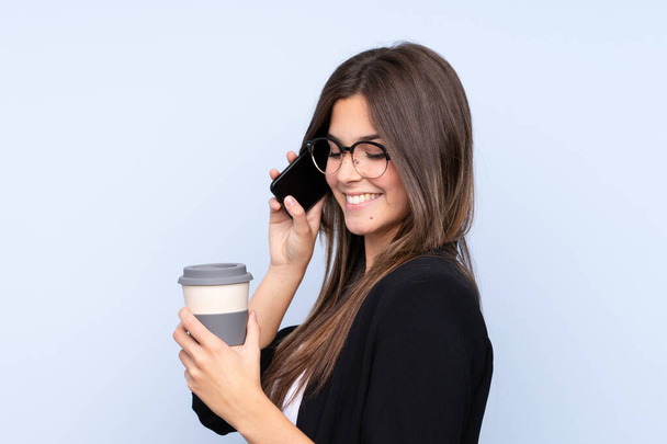 Młoda brazylijska kobieta biznesu trzyma kawę, aby zabrać i rozmawiać z mobile na odizolowanym tle niebieskim - Zdjęcie, obraz