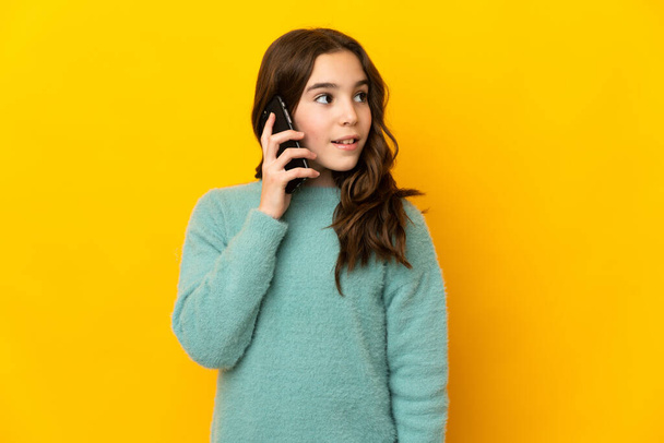 Klein blank meisje geïsoleerd op gele achtergrond houden van een gesprek met de mobiele telefoon met iemand - Foto, afbeelding