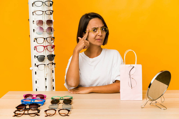 Junge hispanische Frau probiert Brille an, zeigt mit dem Finger auf die Schläfe, denkt nach, konzentriert sich auf eine Aufgabe. - Foto, Bild